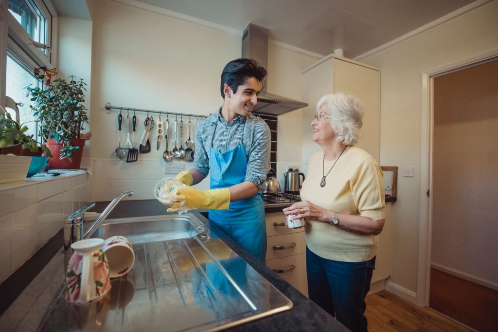 Teenage boy is talking to his grandmother while he washes the dishes in her kitchen.