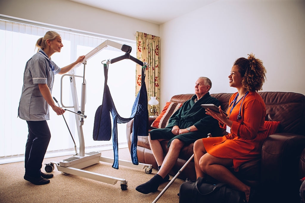 Two homecare nurses at an elderly mans house. They have a hoist set up.